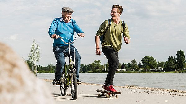 Älterer Mann auf Rad fährt neben jungen Burschen auf dem Skateboard