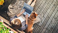 Frau sitz auf ihrem Balkon mit einem Laptop in der Hand. Daneben liegt ein Hund.